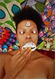 foto de artista deitada sobre tecidos coloridos e com conchas marinhas sobre os olhos e a boca